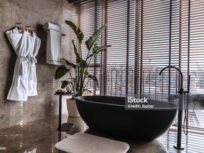 An exclusive penthouse bathroom featuring a sleek, freestanding black tub set against a panoramic city view, framed by large windows with elegant wooden blinds. The harmony of polished marble surfaces, the minimalist black faucet, and the touch of green from a houseplant creates a private urban oasis. This space is the epitome of high-rise luxury, offering a serene escape with the bustling city below. Hanging white bathrobes complete the indulgent, spa-like experience